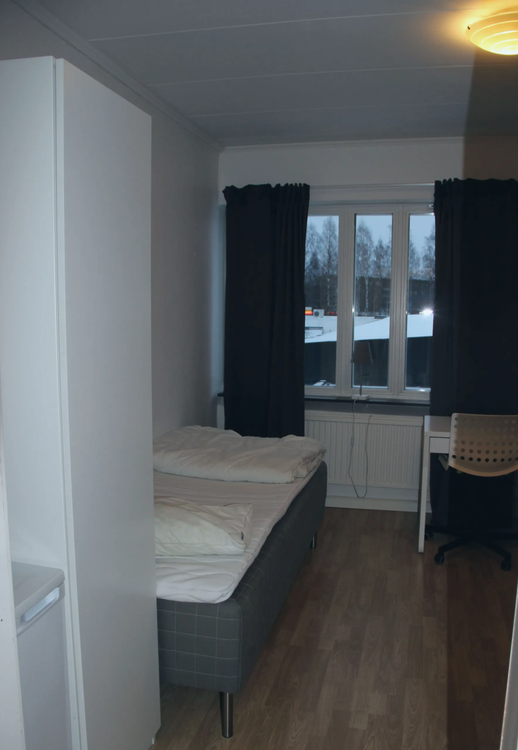 Rum med vita väggar, en säng till vänster ett skrivbord med stol till höger och ett fönster med mörka gardiner på sidorna