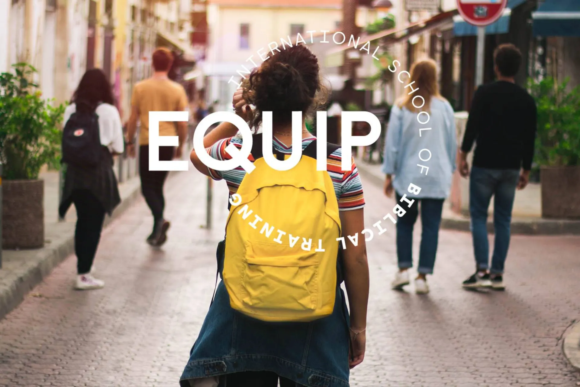 kvinna med ryggen mot kameran. Står på en gata med sex andra vuxna. Kvinnan har en gul ryggsäck på ryggen.