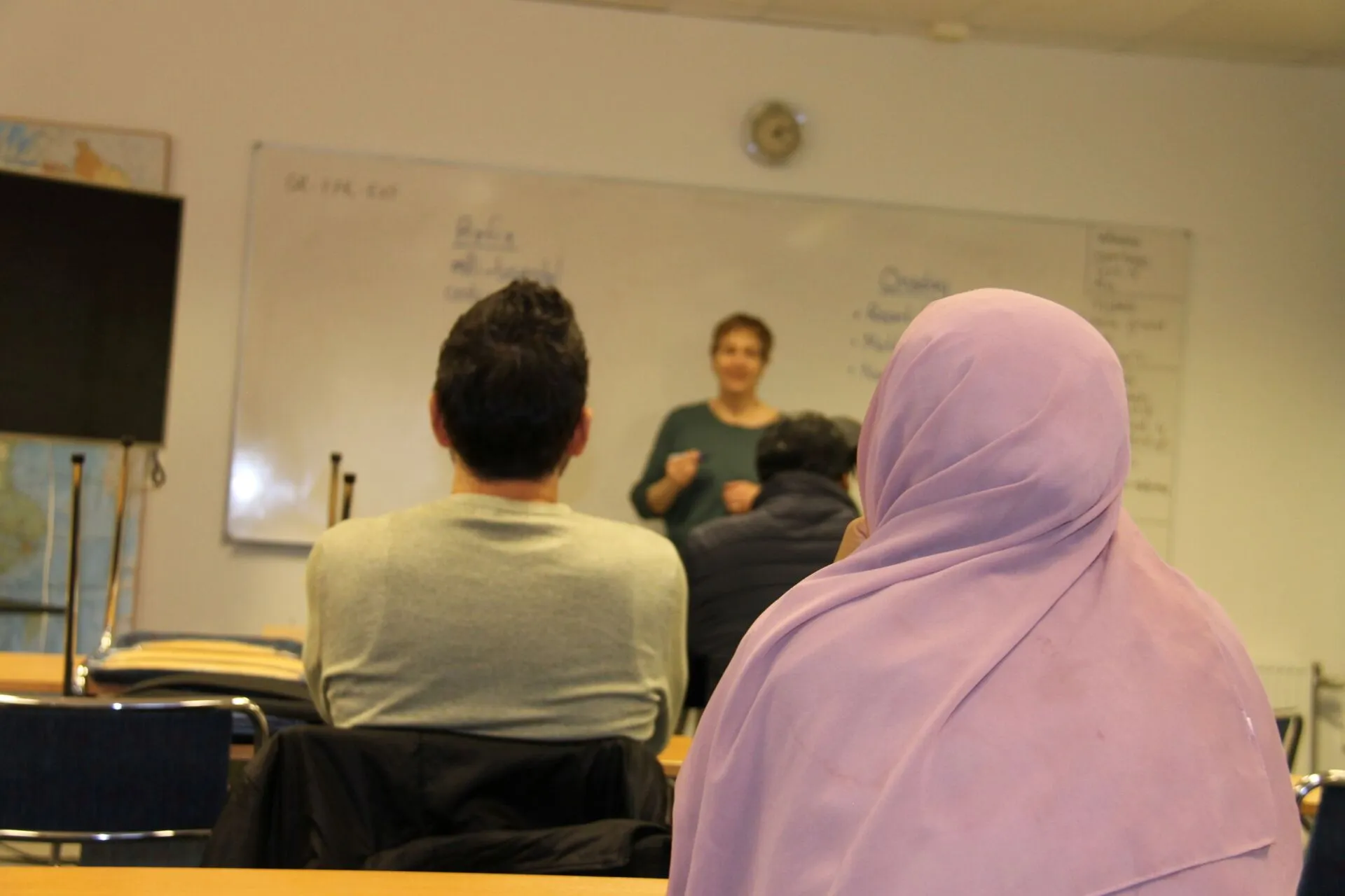 IMG_9295 Klassrum. I bakgrunden står en lärare framför en whiteboardtavla. Till vänster finns ryggen på en man i grå tröja och svart hår, till höger ser man ryggen på en kvinna i lila slöja